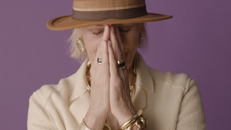 blonde mature woman with blue eyes dressed in jacket, hat and accessories posing with hands on her face and smiling at camera on purple background