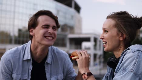 Una-Pareja-Joven-Y-Feliz-Está-Pasando-El-Rato-En-El-Parque.-Riendo-Y-Sonriendo.-La-Chica-Está-Comiendo-Hamburguesa.-El-Joven-Limpia-Suavemente-La-Salsa-De-Los-Labios-De-Su-Chica-Y-Lame-Un-Dedo.-De-Cerca