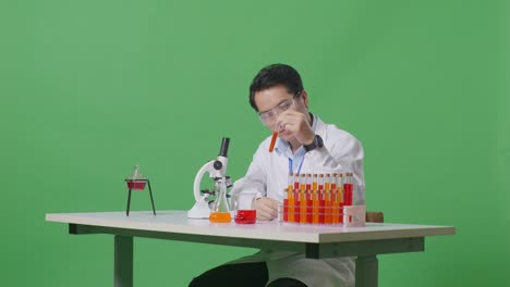 side view of asian man scientist looking at the red liquid in the test tube and shaking head on the table with microscope in the green screen background laboratory
