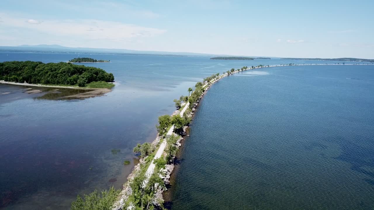 Aerial view rising above a causeway bike path in the north of Lake Champlain during the summer, with clear blue water, green trees, and people exercising.