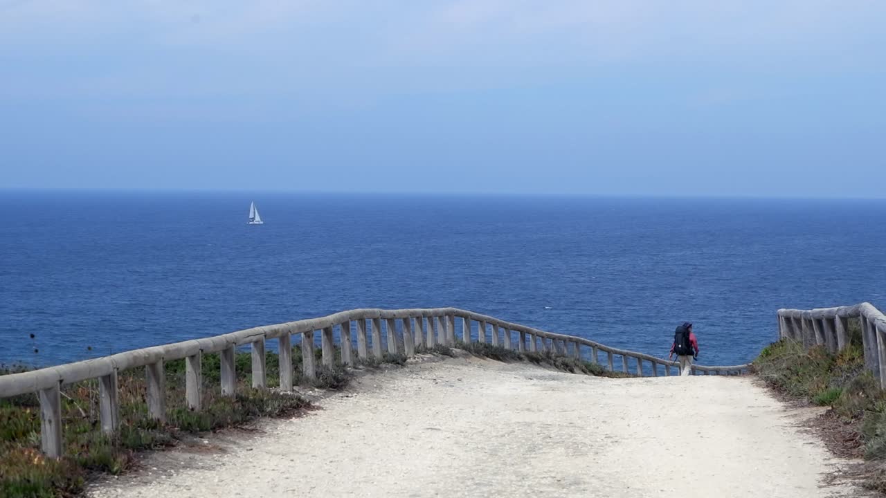 Backpacker hikes dirt road above vast ocean view with one sailboat