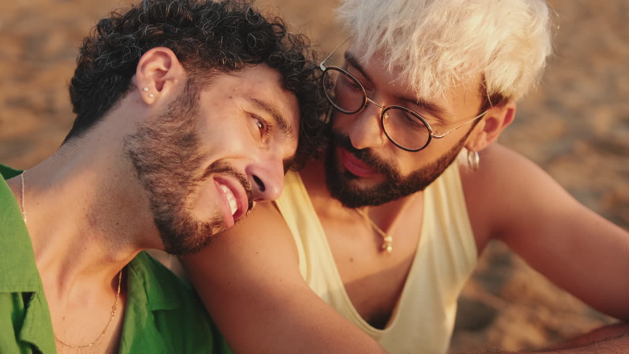 Primer plano de una pareja gay feliz hablando y sonriendo en la playa al atardecer.