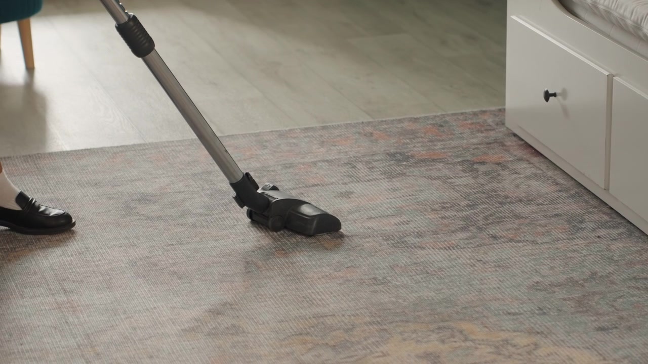 Caucasian woman vacuuming house carpet rug near storage bed, closeup of vacuum nozzle gliding across textured carpet, laminate flooring visible, loafer shoe stepping, smooth backandforth motion