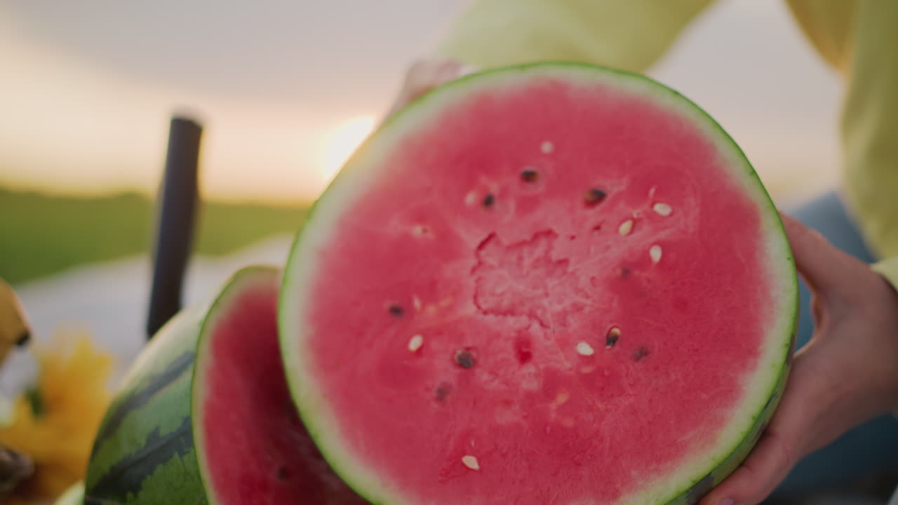 Manos sosteniendo una sandía madura al atardecer, primer plano de una escena de picnic sobre una manta con una cesta de plátanos, luz de la hora dorada, mano revelando la jugosa pulpa roja recién cortada con semillas, ambiente relajado de verano