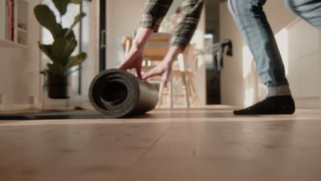 Young-male-folding-yoga-mat-on-the-floor-at-home-in-sunlit-living-room