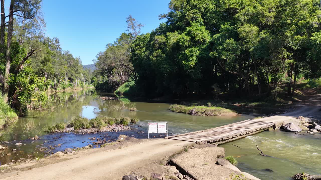 A tranquil forest stream flows under a rustic wooden bridge, surrounded by lush greenery and clear blue skies