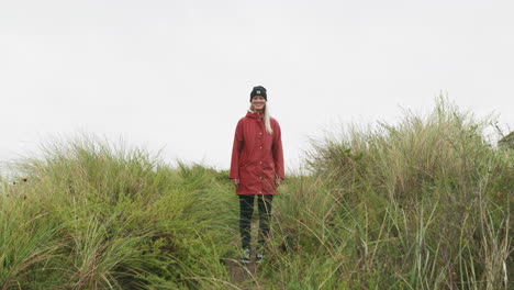 caucasian girl in red jacket standing and walking away by the grassy path towards the ijmuiden beach in the netherlands