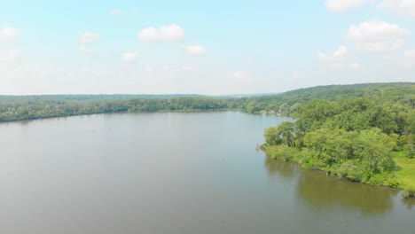 drone-flight-left-over-people-kayaking-on-forest-lake-in-a-forest-preserve-of-green-trees-and-a-blue-and-white-cloudy-sunny-sky-4k