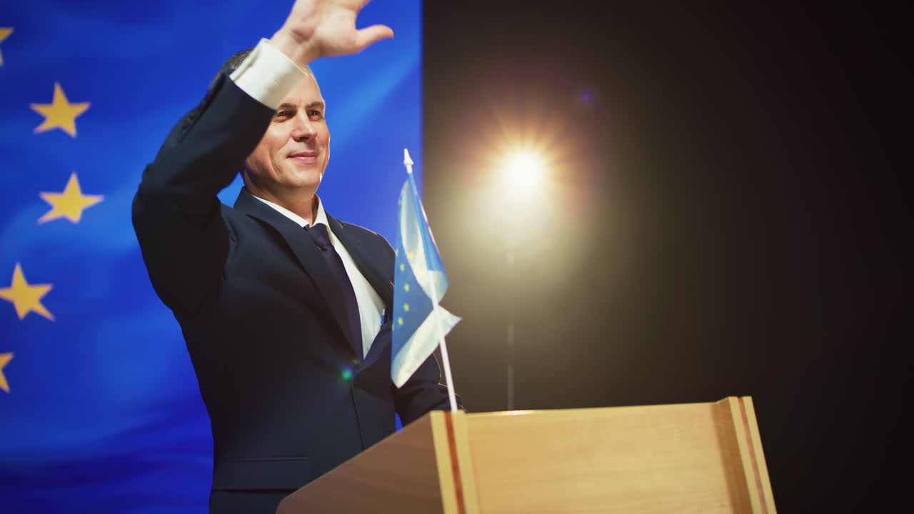 Cheerful Politician Greeting Audience from Podium Zoom out View of Positive Mature Man in Suit Waving Hand and Smiling to Audience while Standing on Rostrum against European Flag against Photo Camera Flashes