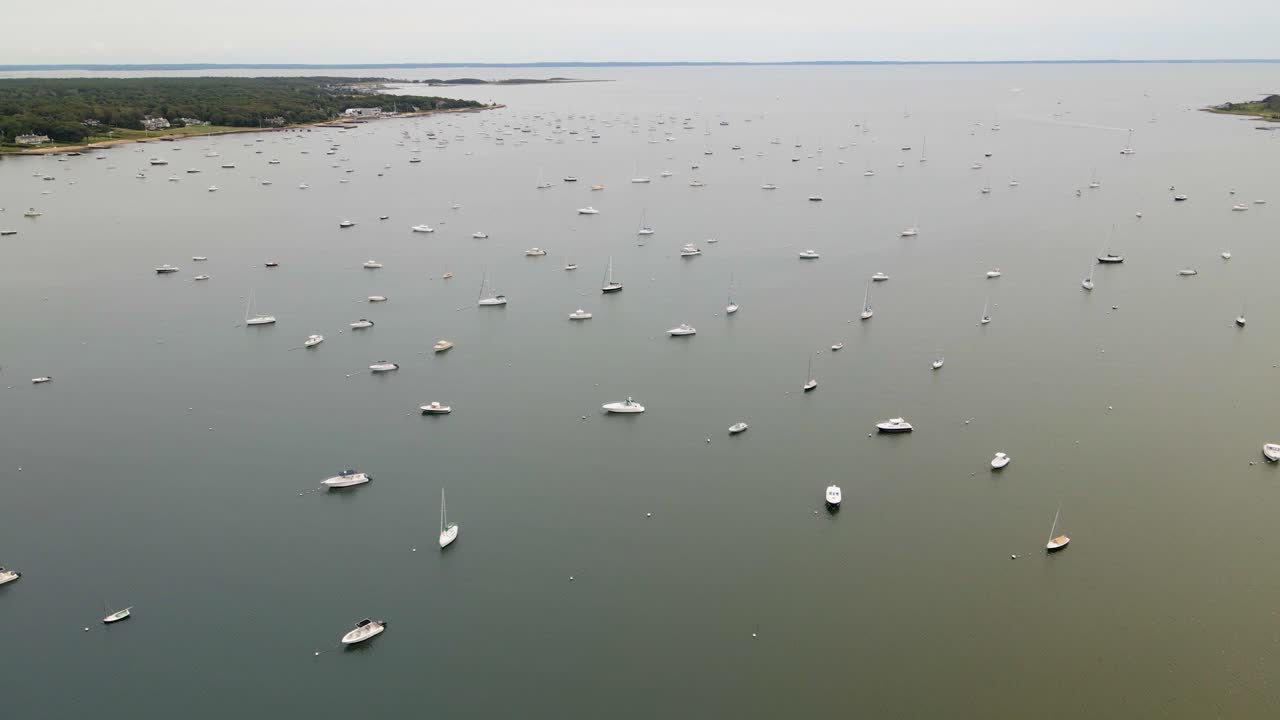 Droneoptagelser viser talrige sejlbåde og yachter fortøjet i en fredelig kysthavn med grøn kystlinje synlig i baggrunden.