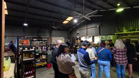 a group of people stand in line inside a rustic farm shop, illuminated by overhead lights, as they wait to purchase local produce in stanthorpe, queensland