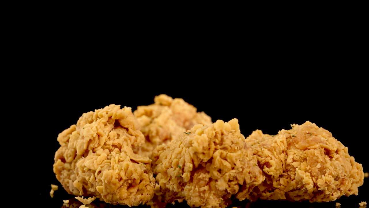 A hand selects a piece of golden, crumbed fried chicken from a pile on a glossy black surface, shot in dramatic slow motion with studio lighting