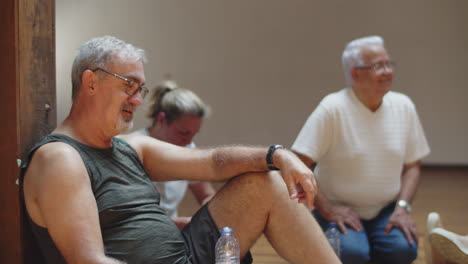 Happy-senior-man-sitting-on-floor-after-dance-class-in-ballroom,-drinking-water-from-plastic-bottle