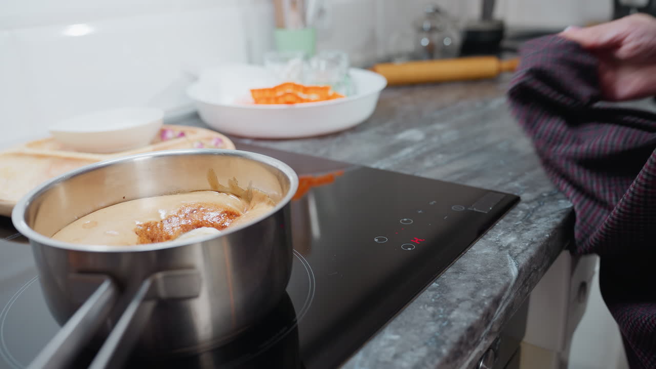 Close-up of food cooking on electric stove with various kitchen utensils nearby as person wipes sleek glass surface with cloth in clean, modern kitchen, smooth countertop and organized workspace