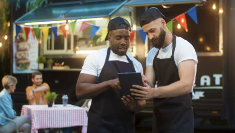 Two-Cheerful-Male-Waiters-In-Aprons-And-Caps-Standing-Outdoor-Foodd-Truck-At-Park-Festival