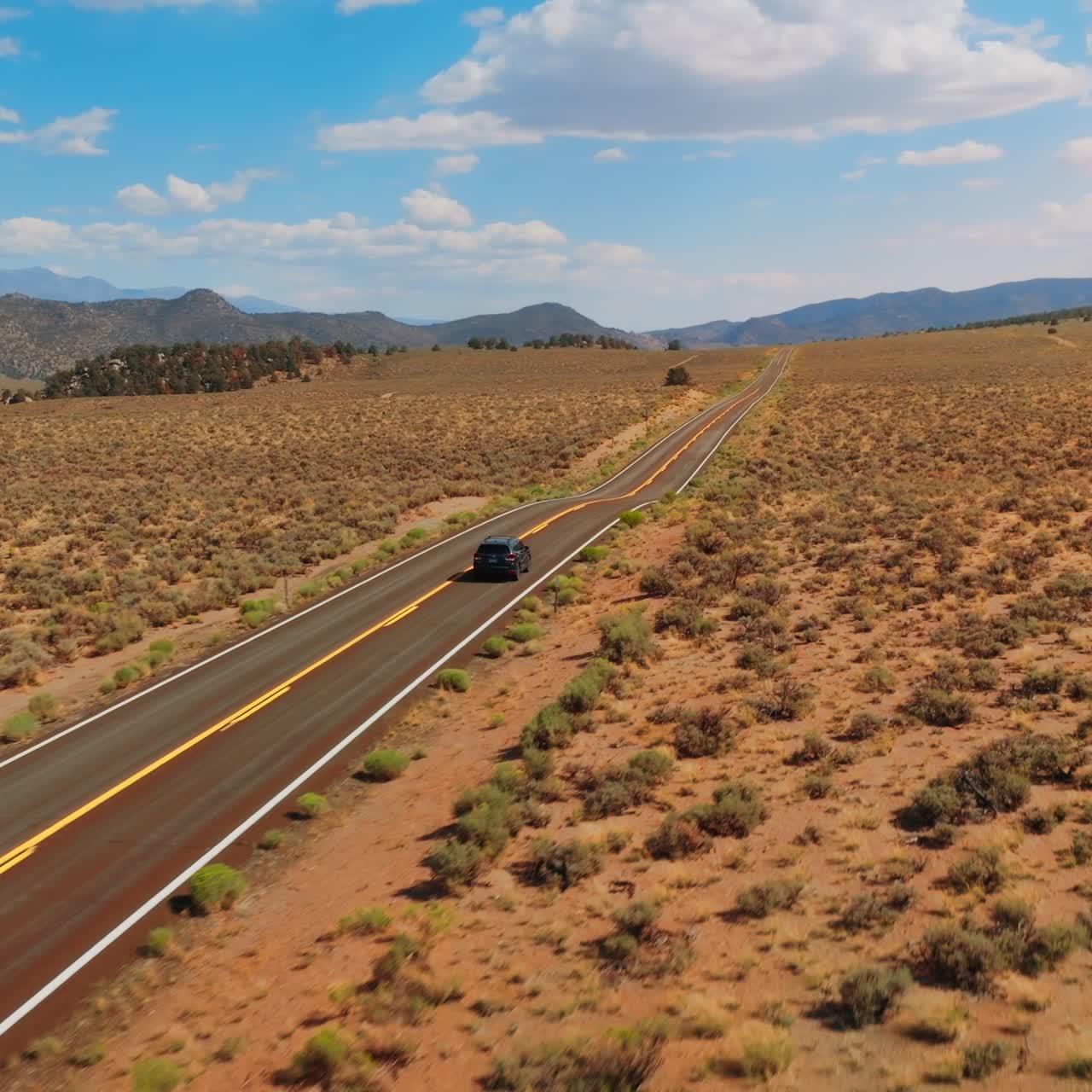 Automobile moving by the two-lane road along the dry sandy landscape. Bare mountains at backdrop of azure sky with fluffy clouds