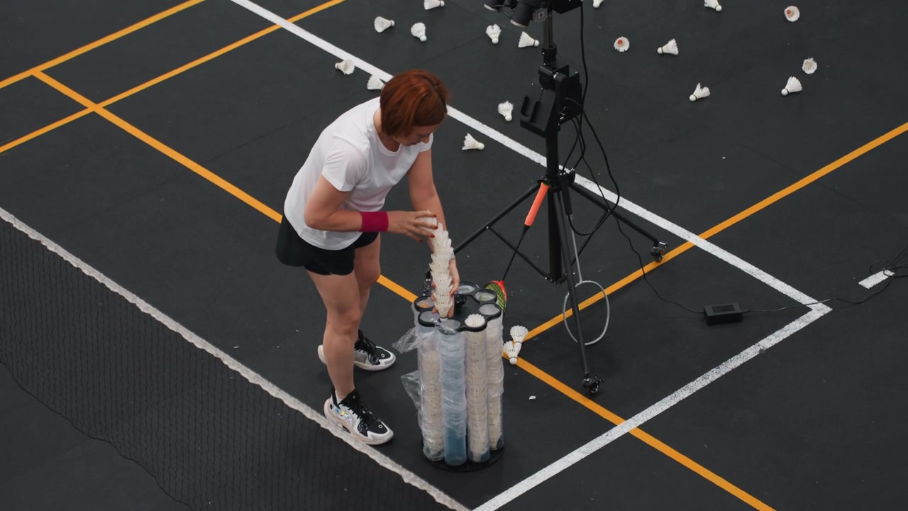 Athlete prepares shuttlecock dispenser on court, overhead shot shows scattered shuttlecocks, tripodmounted camera and training machine, concentrated warmup routine and systematic equipment arrangement