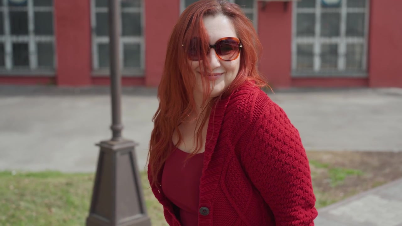 Close up of woman walking outdoors on paved ground holding and swinging maroon knitted fabric. Woman wears red knitwear with dark jeans and white heeled shoes carrying item in her hand