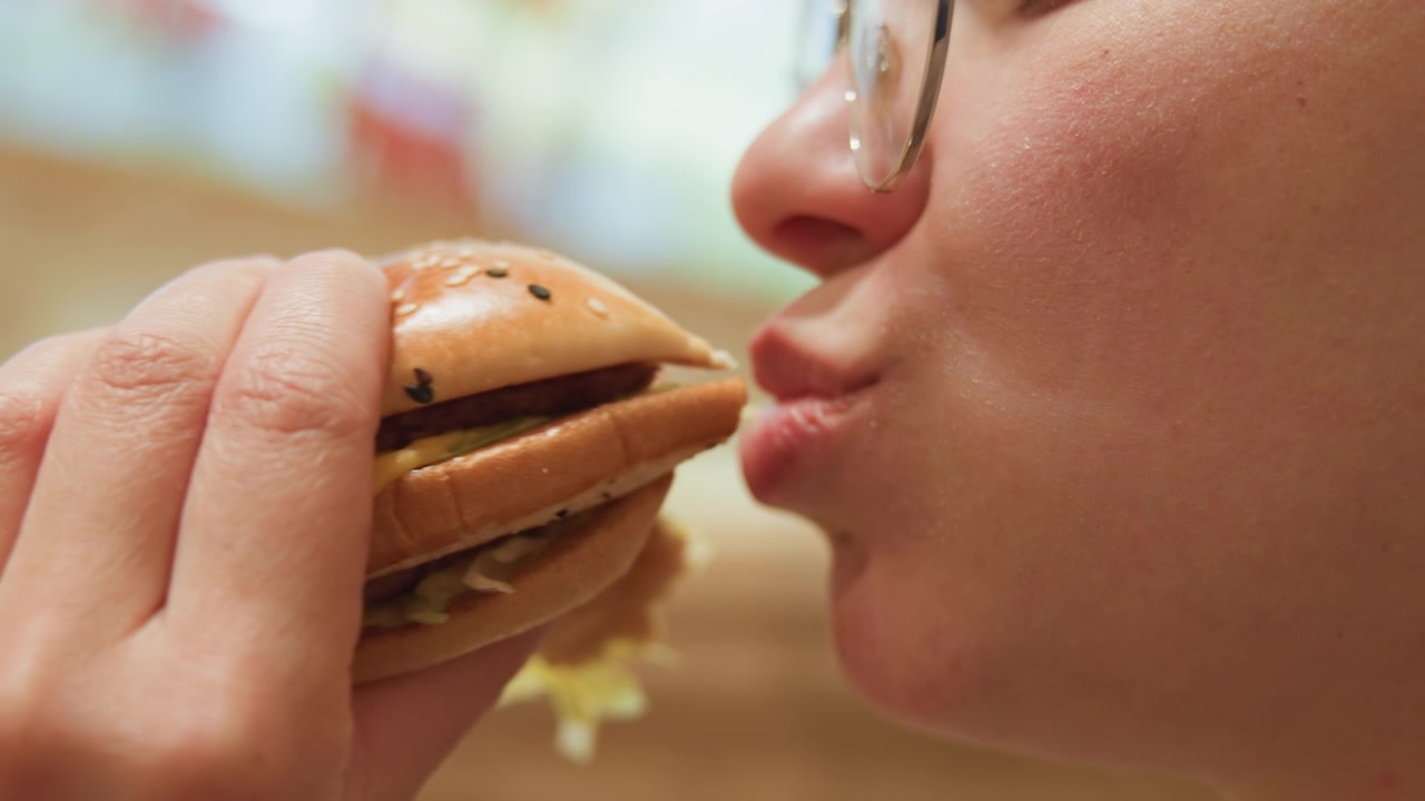 Woman eats burger. Closeup of woman biting cheeseburger. Candid shot of woman savoring savory burger with relish. Close portrait of woman biting into cheeseburger with messy sauce and glasses