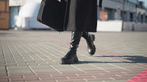 side leg view of person in black boots and coat walking across an interlocked path, featuring parked green bicycles and urban building blur in the background