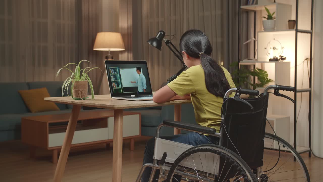 Back View Of Asian Kid Girl Sitting In A Wheelchair While Learning Online With Teacher On Laptop Computer At Home
