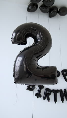 a balloon in the shape of number two. black balloons and happy birthday sign on the white wall. vertical video.