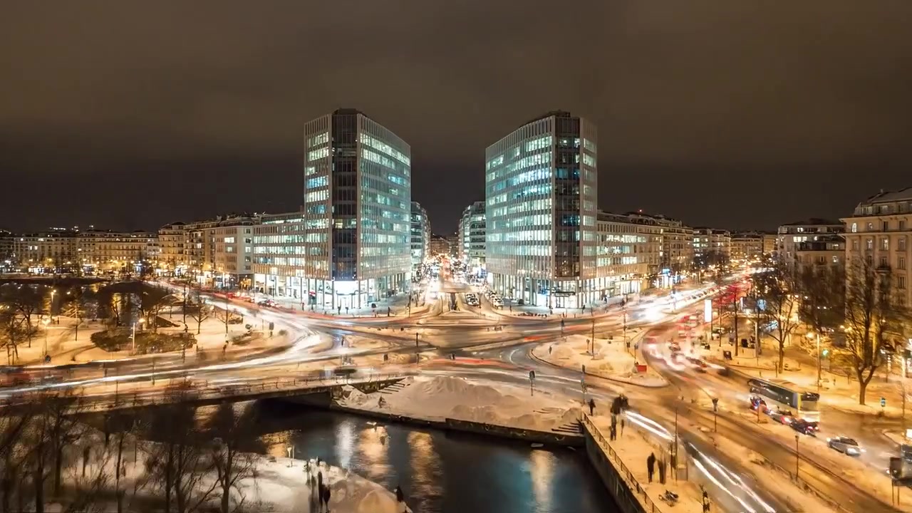 Nattlig byscene med moderne bygninger og travel trafikk midt i et snølandskap, med refleksjoner i vann i et fortryllende bybilde i skumringen.