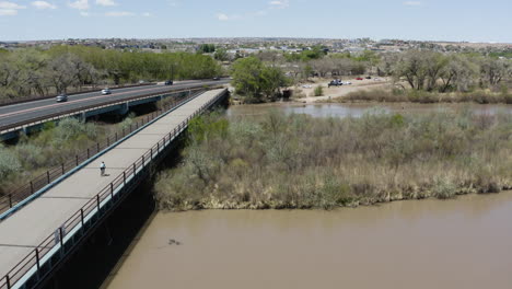 Persona-Montando-En-Bicicleta-En-El-Puente-De-La-Carretera-Sobre-El-Río-Grande-Por-Albuqurque,-Nuevo-México---Antena