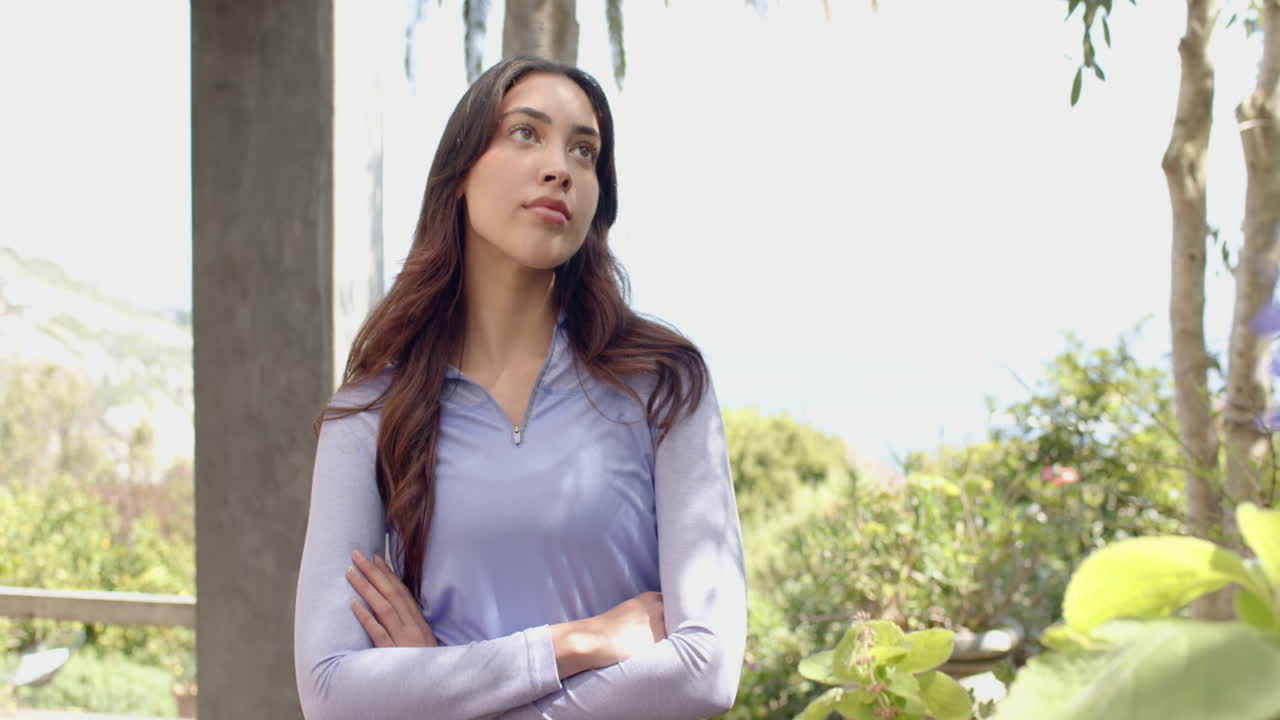 Woman in purple top standing outdoors, arms crossed, looking thoughtful
