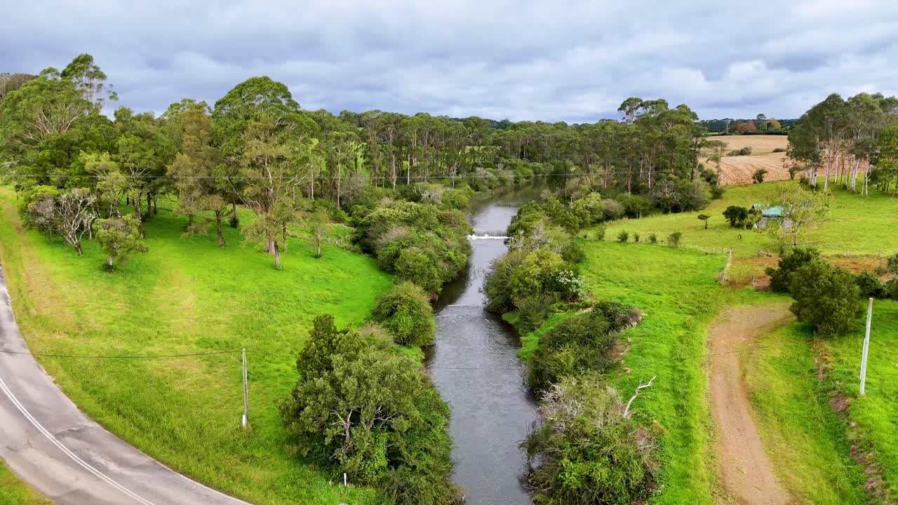 Drone glides above green farmland, winding stream, and trees under cloudy daylight in Dorrigo