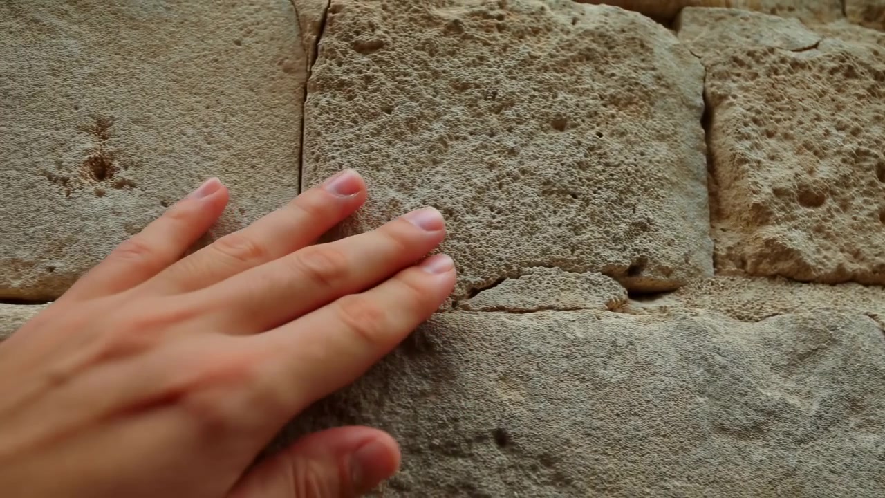 Mano che tocca un antico muro di pietra, primo piano, polpastrelli di persona non specificata che esplorano la muratura ruvida in arenaria