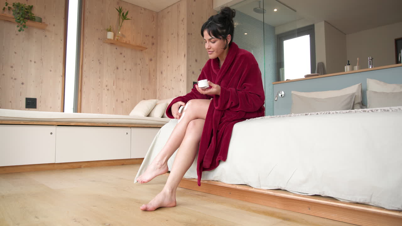Woman in red robe applying lotion on legs, relaxing in cozy bedroom