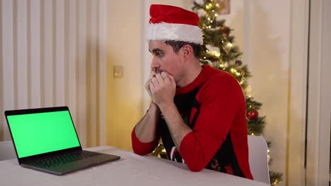 Joven-Asustado-Con-Gorro-De-Papá-Noel-Sentado-En-La-Mesa-Junto-Al-árbol-De-Navidad-Y-Mirando-La-Pantalla-Verde-De-Un-Portátil