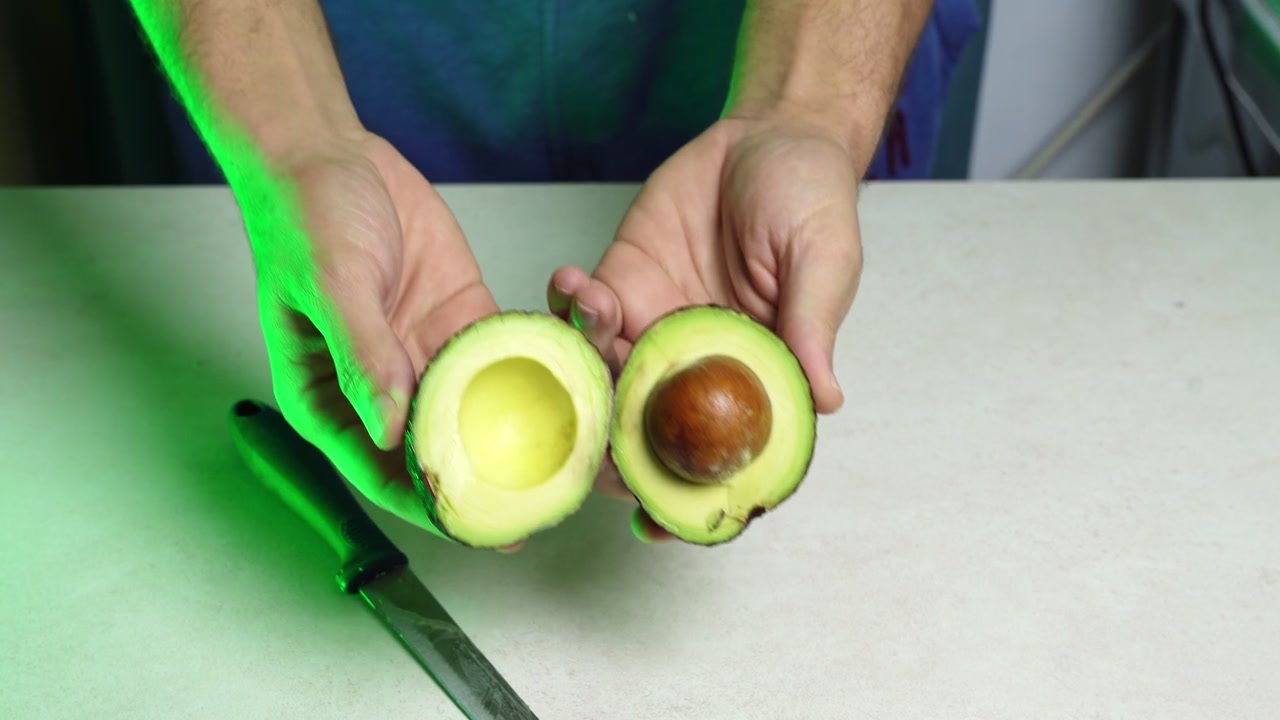 Un primo piano di mani maschili che usano un coltello affilato per tagliare a metà un avocado fresco. Le mani mostrano il frutto aperto rivelando la polpa verde e il nocciolo marrone su un bancone da cucina con illuminazione verde