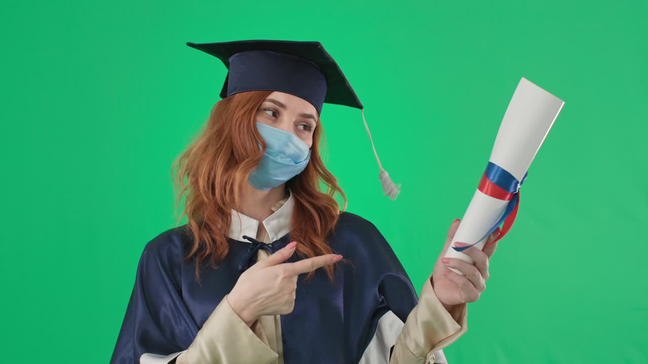 원격 교육, 학술복과 의료 마스크를 입은 여성 졸업생 대학 졸업장을 들고 온라인으로 공부하는 동안 손가락을 내려 놓습니다.