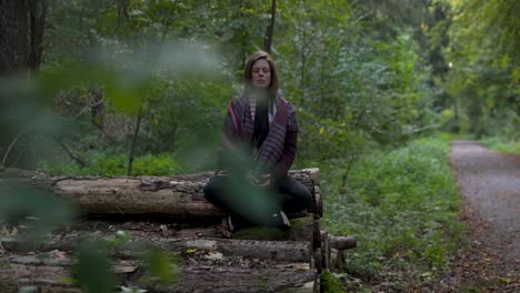 thoughtful natural women meditating in the green forest while sitting on logged trees