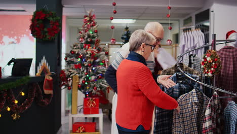 Kamerafahrt-Eines-älteren-Paares,-Das-Während-Des-Weihnachtseinkaufs-In-Einem-Festlich-Geschmückten-Bekleidungsgeschäft-Durch-Hemden-Stöbert.-Älterer-Ehemann-Und-Ehefrau-Auf-Der-Suche-Nach-Formeller-Kleidung