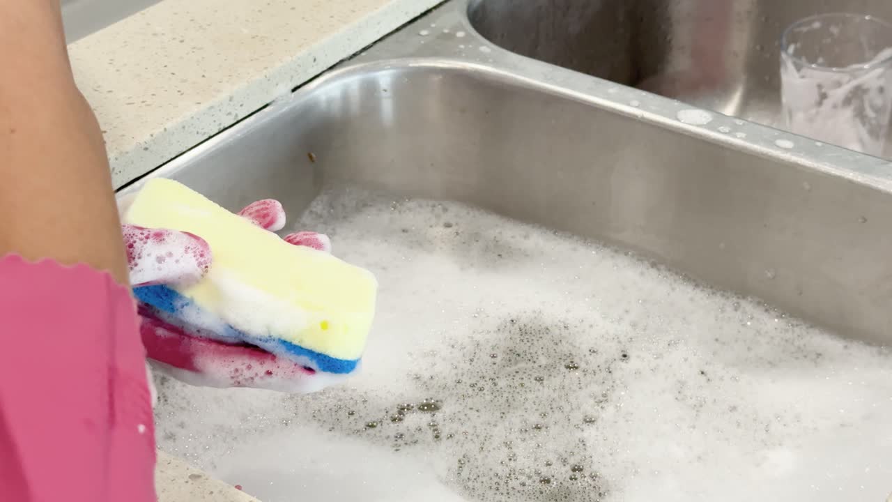 Person wearing pink gloves scrubs and rinses dirty plates with a sponge in a well-lit kitchen sink, using soapy water and thorough cleaning motions