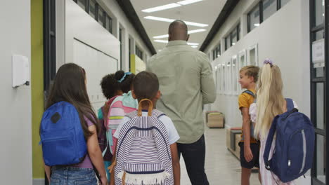 video of back of african american male teacher and diverse pupils walking at school hall