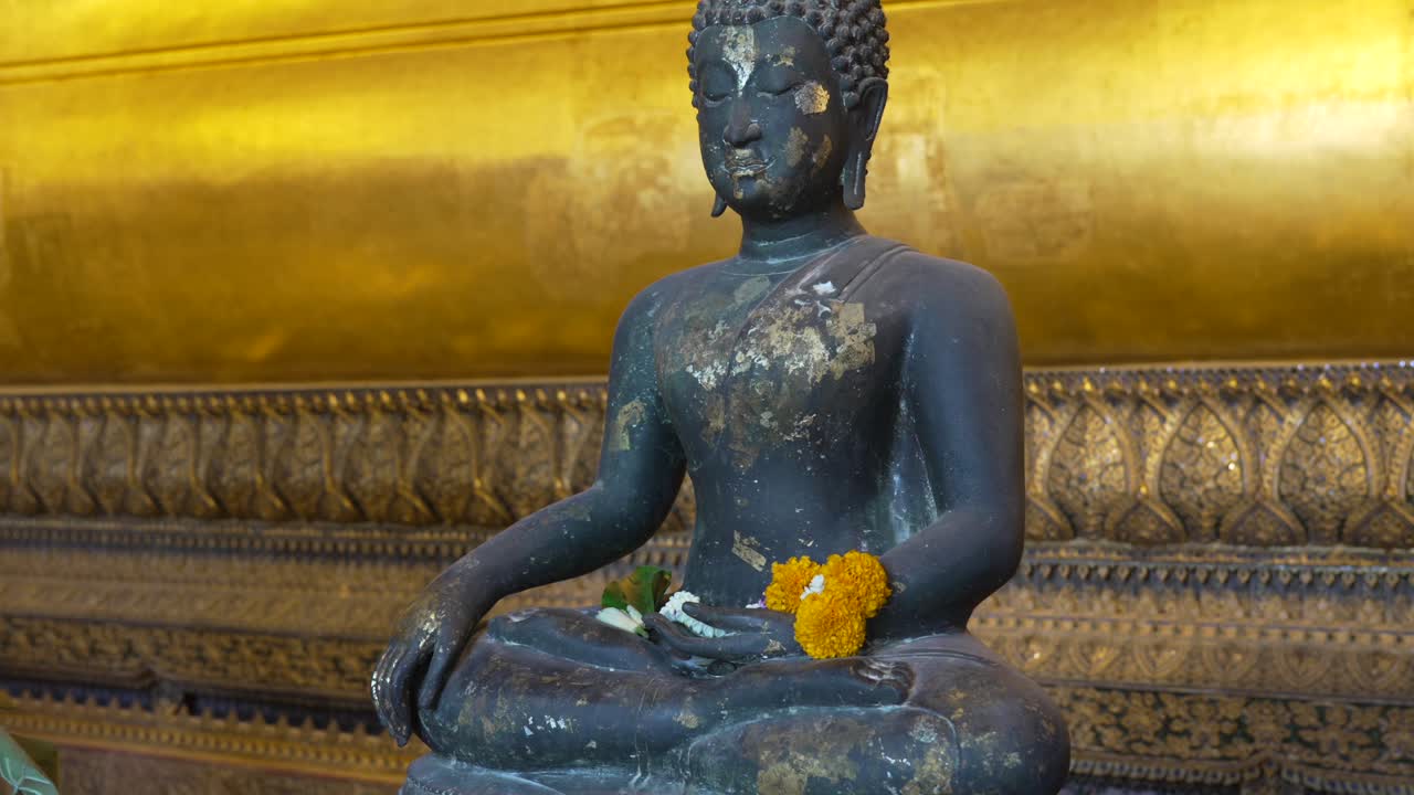 Seated dark Buddha statue displaying golden leaf patches and a marigold garland, embodying devotion and spiritual serenity within the sacred Wat Pho temple in Bangkok, Thailand