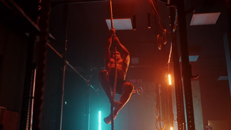 fitness man doing rope climb exercise in gym slow motion. orange blue neon light. man working out in a gym climbing a rope