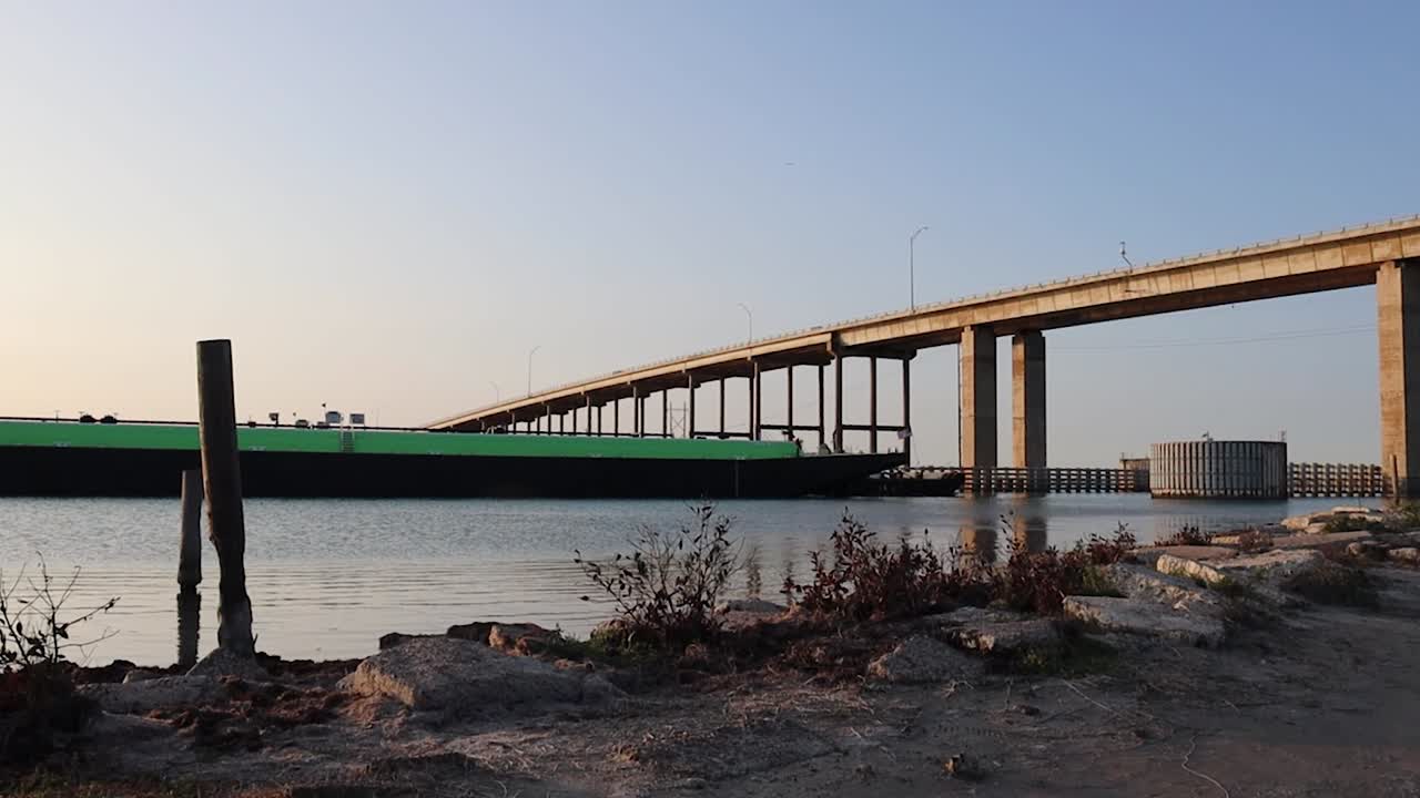 대형 녹색 바지선이 인터코스탈 수로를 따라 이동하고 석양에 jfk 메모리얼 코즈웨이 다리 아래에서 활공하는 모습을 지켜보고 있습니다.