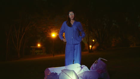 mujer modelo de pie bolsas de basura después de la limpieza parque nocturno bajo linternas ambientales.