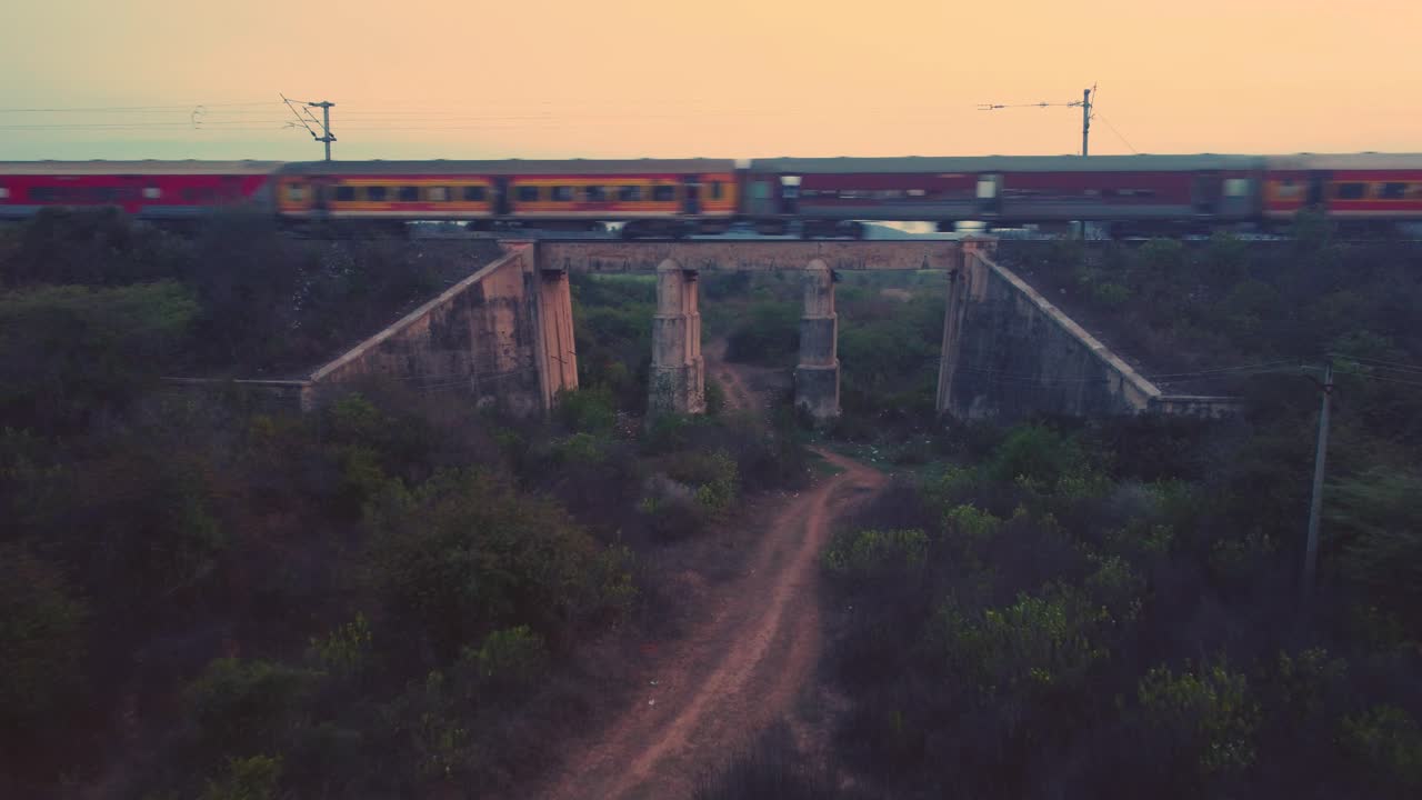 tomada aérea de un tren de pasajeros de los ferrocarriles indios que se mueve rápidamente en un viejo puente ferroviario de hormigón con densas colinas forestales en el fondo durante la tarde
