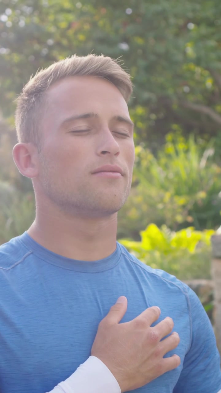 Video verticale: Uomo che respira, con maglietta blu e maniche bianche, mani sul petto, che si calma in giardino, la telecamera si allontana.