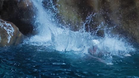 Woman-jumping-off-the-rocks-into-a-natural-pool-of-turquoise-water-surrounded-by-rocks-and-waterfall