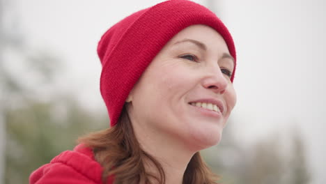 close-up of woman in red beanie and hoodie gazing warmly into distance with soft smile in serene misty setting, capturing introspective and peaceful mood against blurred atmospheric background