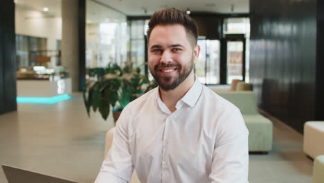 portrait of smiling young businessman using laptop and looking at camera sitting in hotel lobby