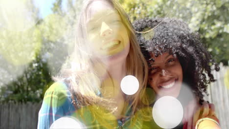 animation of yellow and white light spots over two happy diverse teenage girls embracing in garden