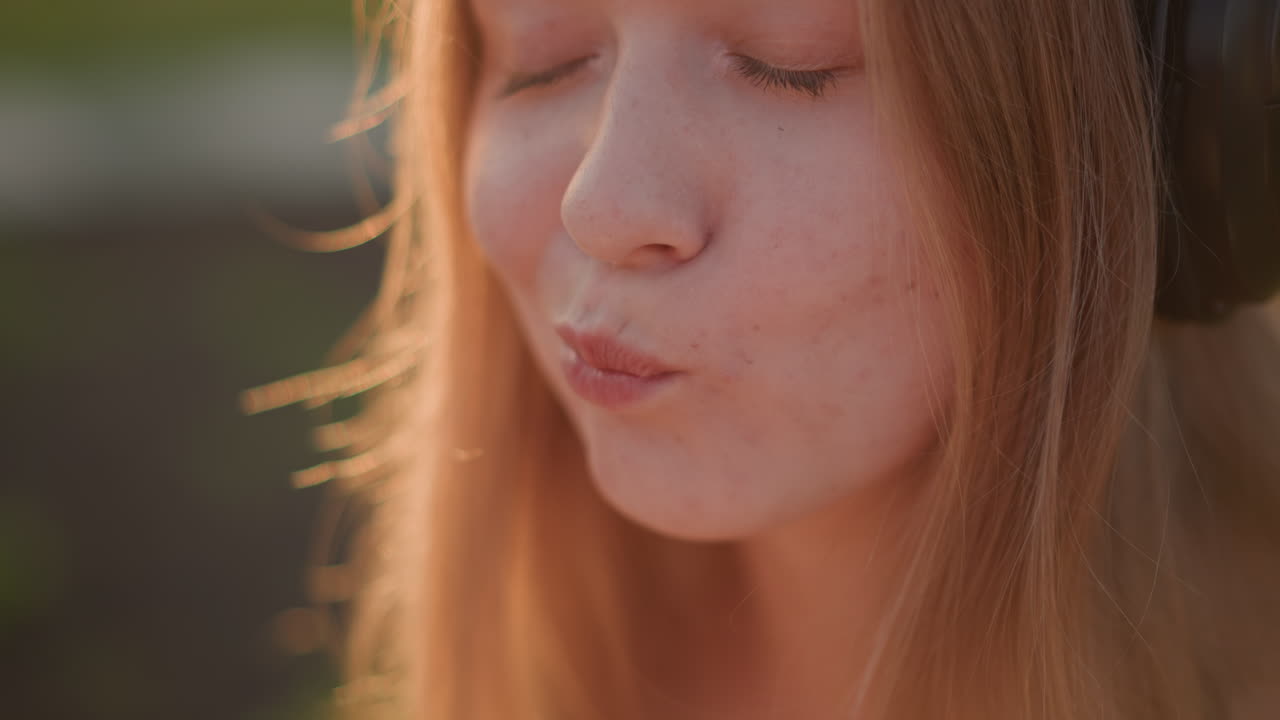 caucasian woman headphones savoring sunset snack, closeup portrait, young woman with freckles, eyes closed enjoying bite, listening to mellow music during golden hour, soft bokeh background, warm